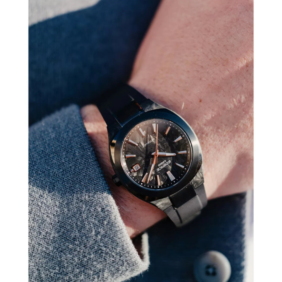 A close-up lifestyle shot of a men's watch on a wrist, featuring a dark dial, black bezel, and a textured gray metal link bracelet, worn over a gray knit sweater.